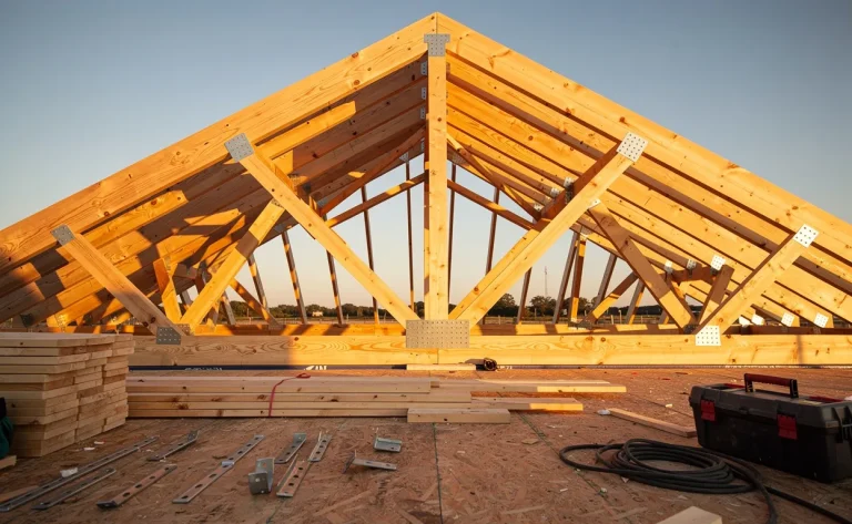 Charpente en bois d’un toit à pignon partiellement construite avec poutres et chevrons, entourée de matériaux de chantier sous une lumière dorée.